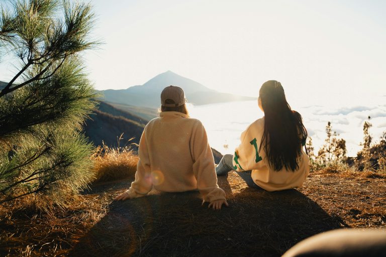 Two people sitting on a hilltop, enjoying a sunset view overlooking scenic mountains and clouds