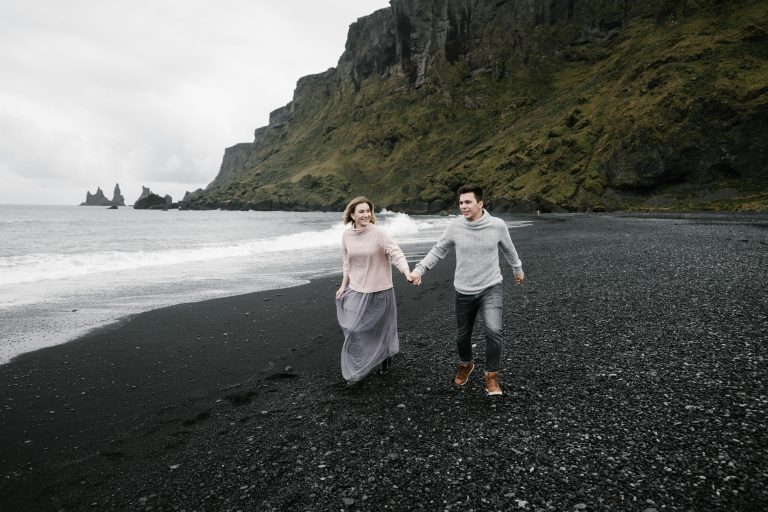 Happy loving couple holding hands and exploring magnificent black gravel beach while enjoying weekend at cliffy seacoast